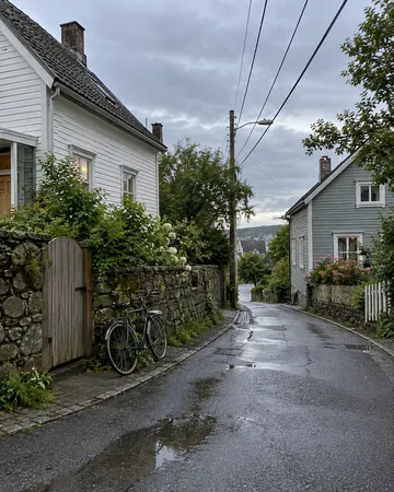 This is a quiet, wet rural street in what appears to be Scandinavia, lined with traditional white wooden houses and stone walls, with no visible people. Notable features include a bicycle leaned against a wooden gate on the left, blooming white flowers, and mountains visible in the misty distance across a body of water. The overcast sky and wet pavement create a peaceful, melancholic atmosphere typical of Nordic countryside.