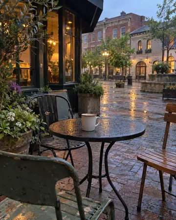 This is an atmospheric evening scene of a wet, rain-soaked European town square or plaza, photographed from a café or restaurant seating area. The foreground shows a small black round table with a white enamel mug, surrounded by metal chairs and potted flowers, while historic brick and stone buildings with illuminated windows line the square in the background. The scene appears deserted of people, creating a quiet, contemplative mood enhanced by the wet pavement reflecting the golden evening light and the cozy glow emanating from the storefronts and building windows.