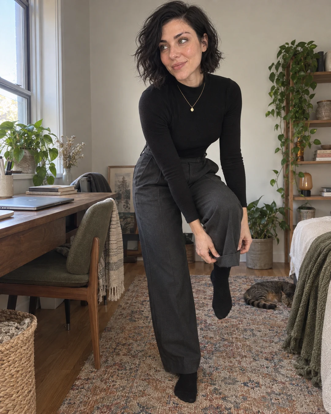 The image shows one person in a cozy home interior, wearing an all-black outfit consisting of a long-sleeve top and wide-leg trousers with black boots, accessorized with a delicate gold necklace. The setting is a well-decorated room with natural wood furniture, potted plants, shelving with books and decorative items, and a warm, minimalist aesthetic with neutral tones. The mood is casual and relaxed, with the person posing in a contemplative manner while a tabby cat rests on the rug in the background.