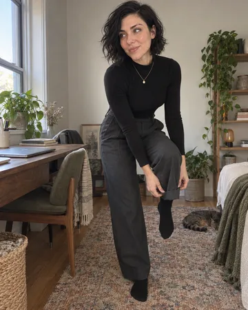 The image shows one person in a cozy home interior, wearing an all-black outfit consisting of a long-sleeve top and wide-leg trousers with black boots, accessorized with a delicate gold necklace. The setting is a well-decorated room with natural wood furniture, potted plants, shelving with books and decorative items, and a warm, minimalist aesthetic with neutral tones. The mood is casual and relaxed, with the person posing in a contemplative manner while a tabby cat rests on the rug in the background.